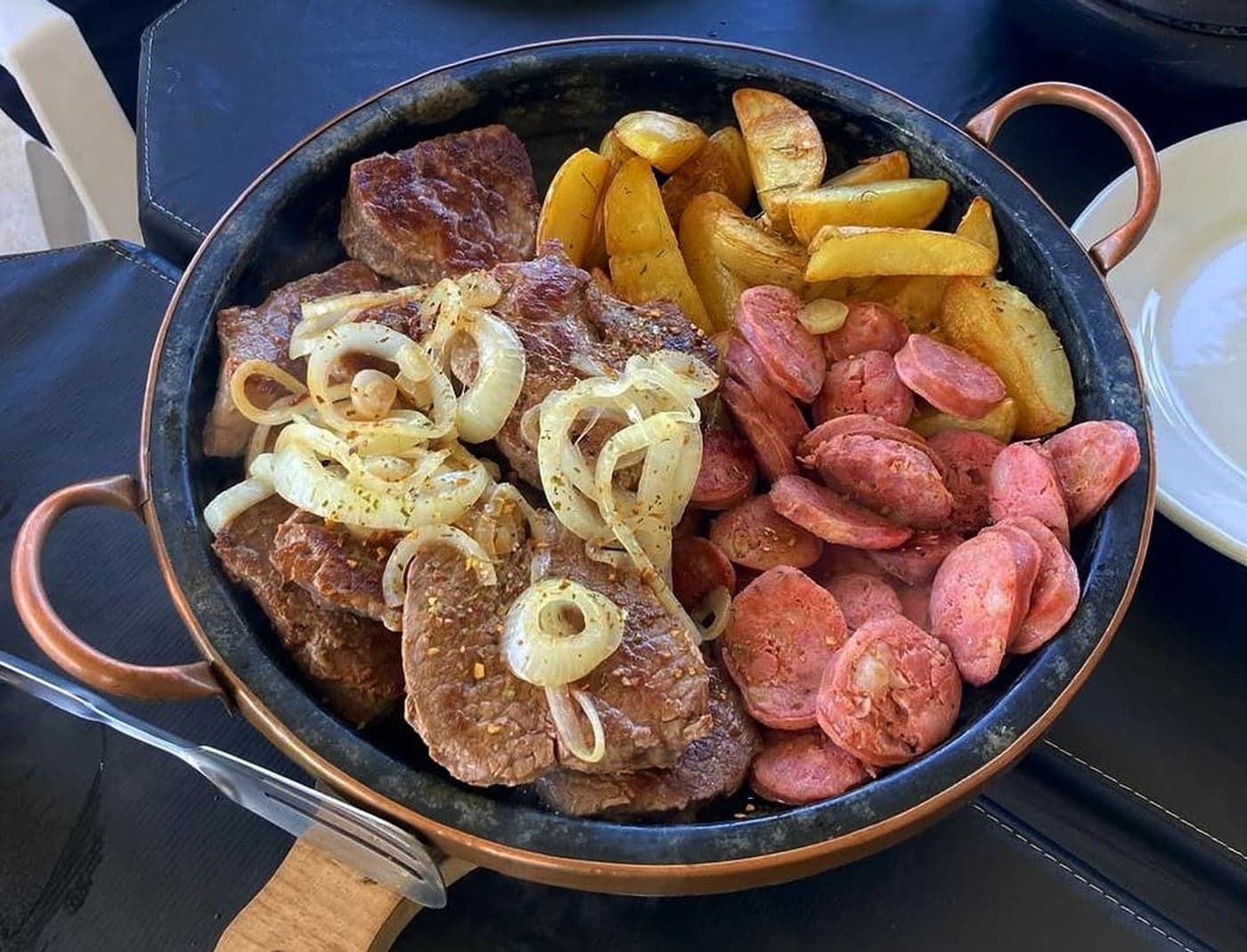 Panela de ferro com bifes, linguiça, banana e cebola servida na mesa do Rancho Aroeira