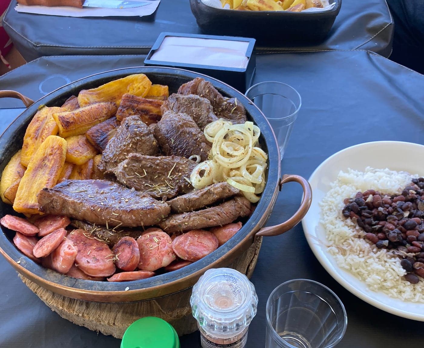Panela de cobre com bifes, banana, linguiça e cebola, ao lado do prato de arroz e feijão